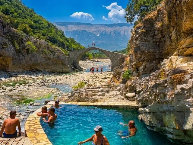 Persone che si rilassano in una piscina termale con un vecchio ponte in pietra sullo sfondo.